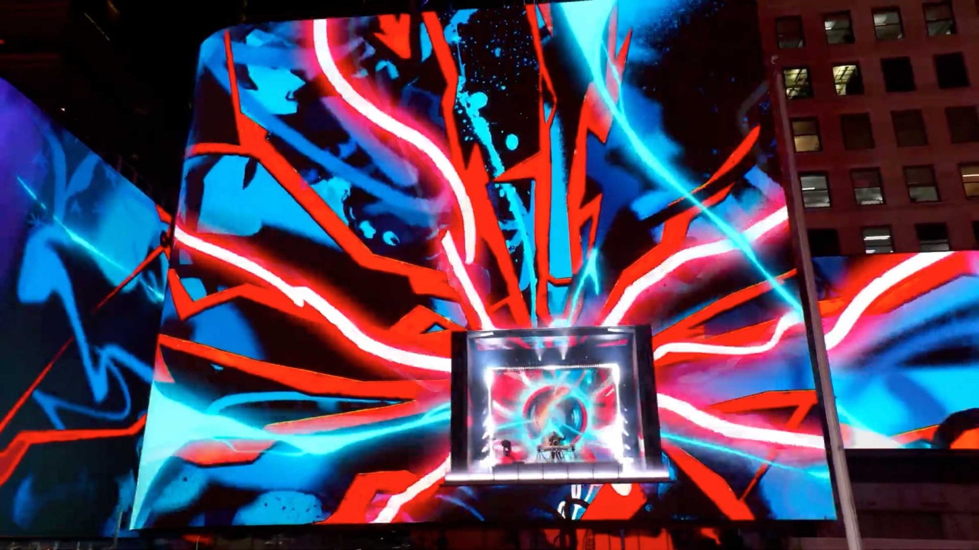 A vivid digital art display on a building in Times Square features bright red, blue, and black abstract shapes radiating from a central screen, creating an energetic and dynamic visual effect at night. Frame from Stash Magazine article.