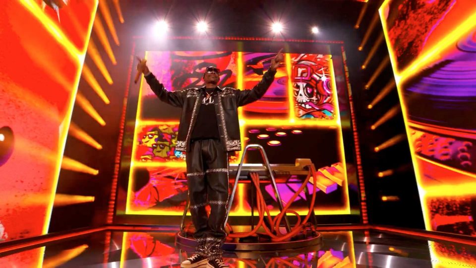 A performer in sunglasses and a black outfit stands on stage, arms raised, as vibrant orange and yellow LED screens display abstract graphics—evoking the energy of Golden Wolf Takes Over Times Square for Epic Games Fortnite. Frame from Stash Magazine article.
