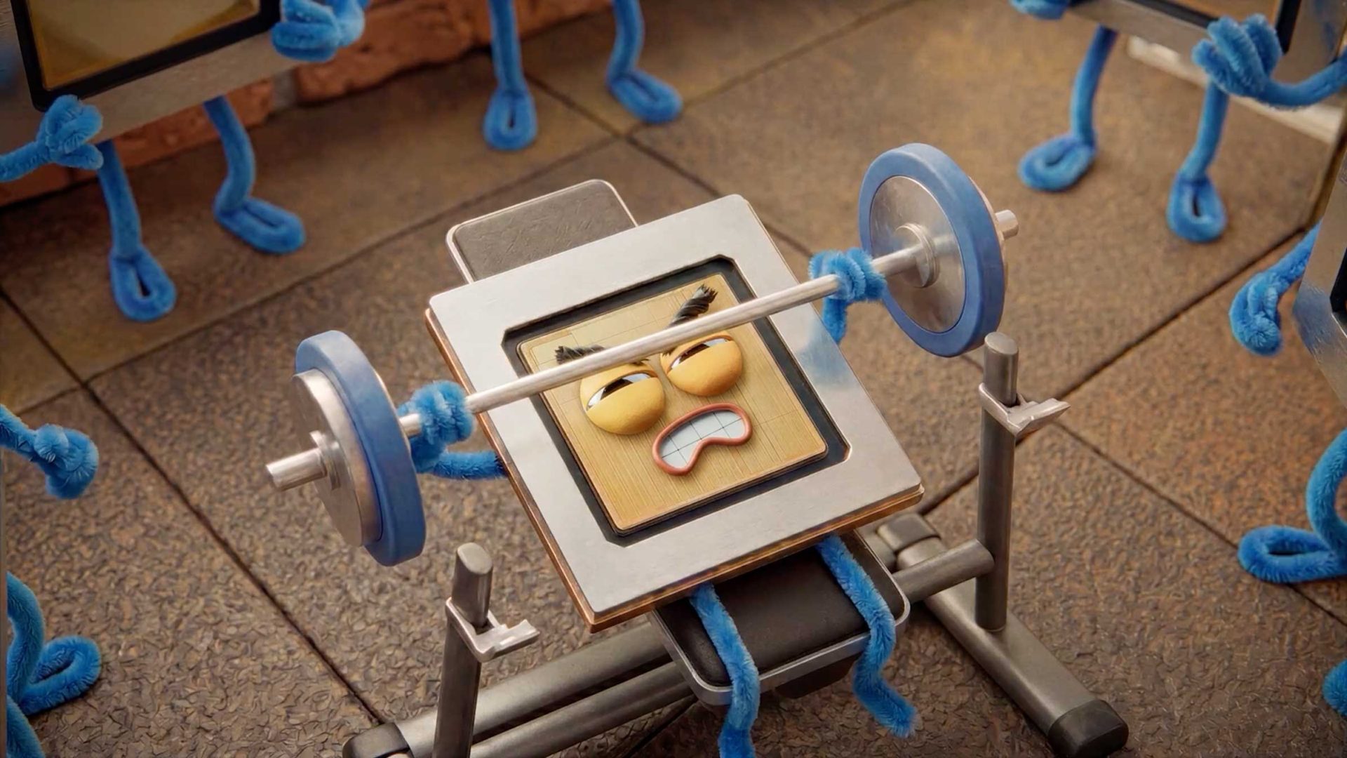 A cartoon tablet with a strained face and blue pipe cleaner arms struggles to lift a barbell on a bench press, surrounded by similar tablet characters in a gym setting—a whimsical scene inspired by the Google I/O Keynote Film 2026 by Laurie Rowan. Frame from Stash Magazine article.