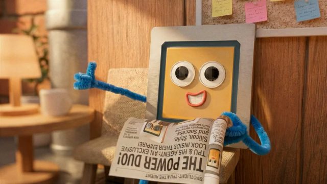 A cheerful, animated square character with googly eyes and blue pipe cleaner arms sits on a chair, holding a newspaper and giving a thumbs-up—ready to tune into the Google I/O Keynote Film 2026 by Laurie Rowan & Nexus in the cozy, note-filled room. Frame from Stash Magazine article.