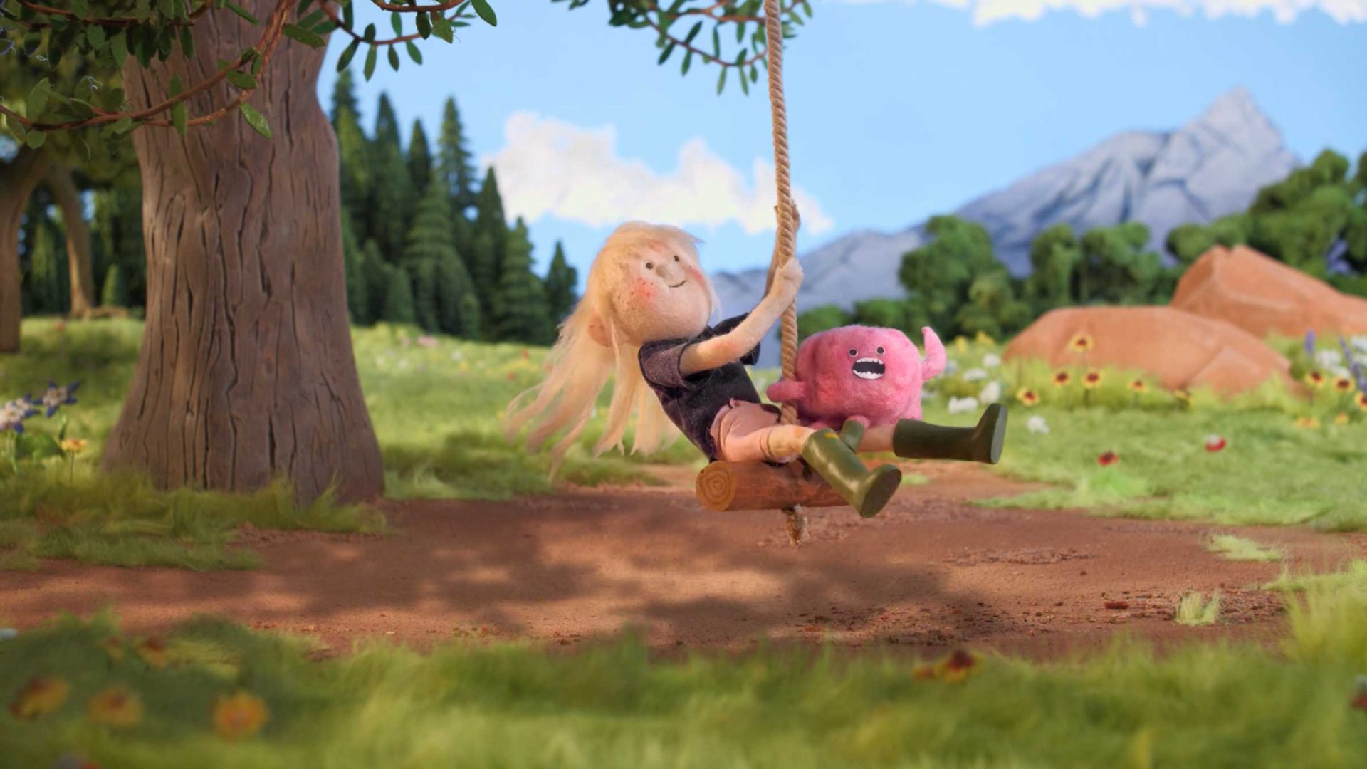 A blonde-haired puppet child swings on a rope swing with a pink stuffed animal in a colorful, lush outdoor setting, celebrating the Great Outdoors Colorado—surrounded by trees, grass, and distant mountains under a blue sky. Frame from Stash Magazine article.