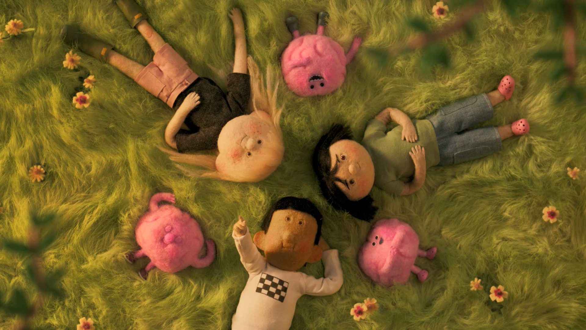 Three kids lie on green grass in a circle, surrounded by four small, pink, plush creatures. Yellow flowers are scattered around as tree branches frame the scene from above. The whimsical and peaceful atmosphere celebrates the wonder of the great outdoors. Frame from Stash Magazine article.