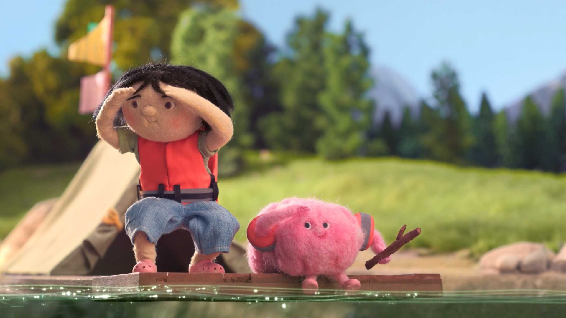 A childlike puppet wearing a red vest and blue shorts sits on a wooden raft next to a pink, fluffy creature holding a stick. Behind them, at their grassy campsite, the spirit of Great Outdoors Colorado and Anna Mantzaris’s whimsy shines through. Frame from Stash Magazine article.