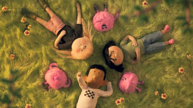 Four animated children lie in a circle on grassy ground, surrounded by small, pink creatures with ears and tails. Flowers dot the scene, creating a peaceful, whimsical mood inspired by Great Outdoors Colorado ad “Take Your Brain Out” by Anna Mantzaris. Frame from Stash Magazine article.