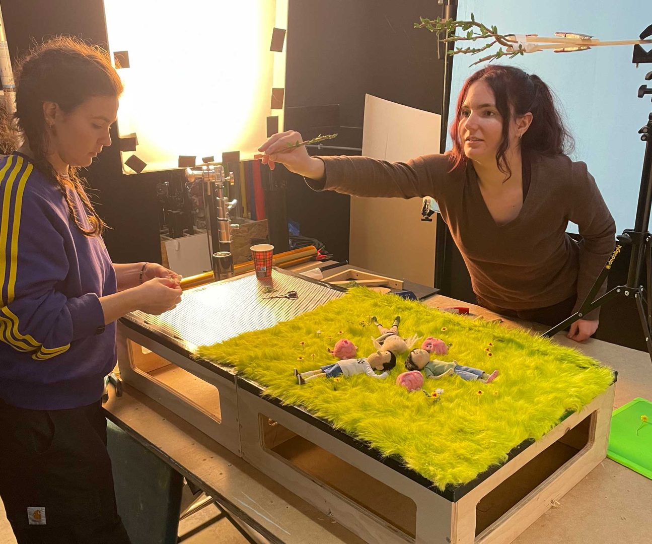 Two people are working on a stop-motion set with a grassy green surface and small character models, channeling Anna Mantzaris’ style. Bright lights and camera equipment surround their workspace as they adjust the outdoors-inspired scene. Frame from Stash Magazine article.