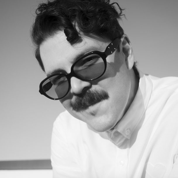 A man with short, wavy hair and a thick mustache wears dark, oversized glasses and a white button-up shirt. In this black-and-white photo, he looks at the camera with a slight smile—ready to Get Seen at OFFF 2026. Frame from Stash Magazine article.