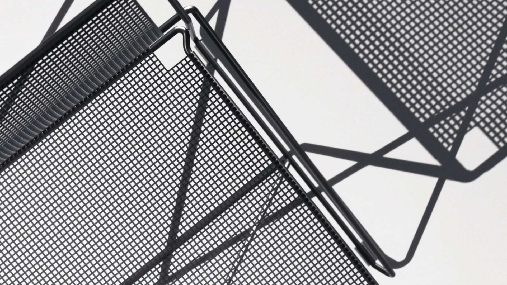 Close-up of black metal mesh outdoor chairs from HAY casting geometric shadows on a white surface, creating an abstract pattern with intersecting lines and shapes. Frame from Stash Magazine article.