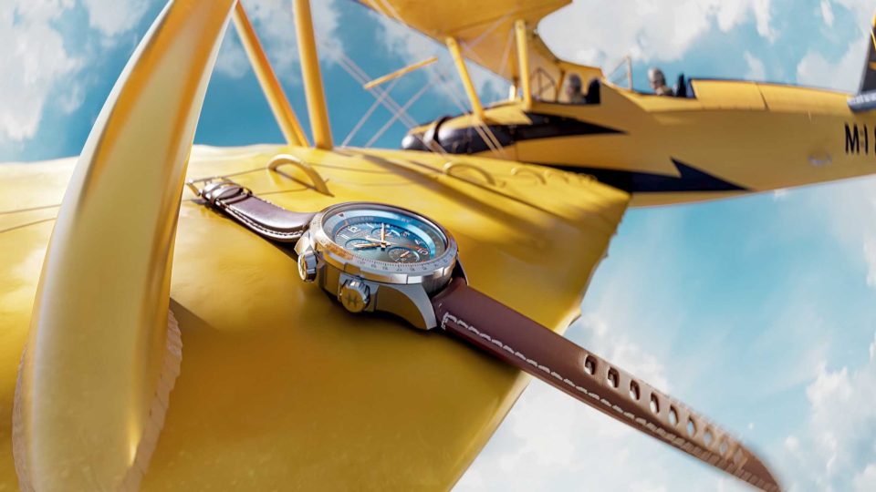 A wristwatch with a brown leather strap rests on the bright yellow wing of a vintage biplane, evoking the adventurous spirit seen in the Hamilton Watches in Action Brand Film by Kutuko, with blue skies and clouds completing the scene. Frame from Stash Magazine article.