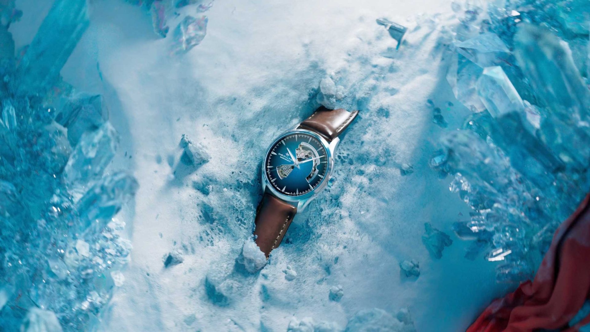 A Hamilton wristwatch with a blue face and brown leather strap is partially buried in snow, surrounded by clear blue ice crystals, evoking the cool, cinematic mood of a Kutuko brand film. Frame from Stash Magazine article.