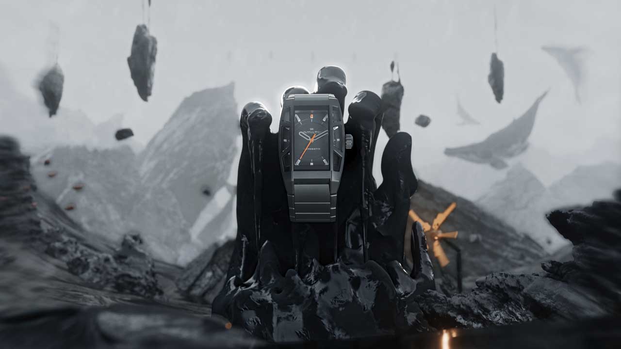A sleek black wristwatch with an orange second hand is displayed upright, surrounded by glossy, black liquid shapes. Inspired by the Hamilton Watches in Action Brand Film by Kutuko, floating rocks and a misty, mountainous landscape appear in the surreal background. Frame from Stash Magazine article.