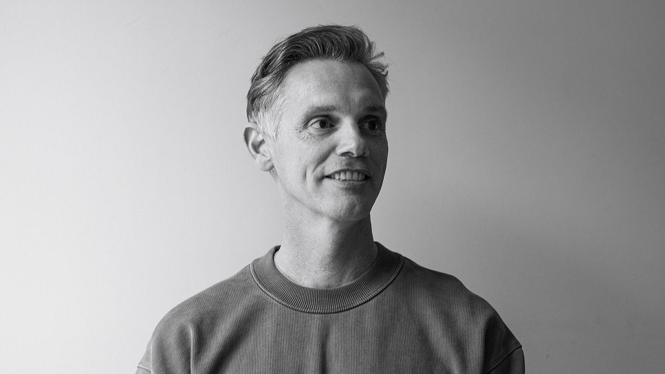 A black-and-white photo of Adrian Lawrence, Heckler's newly appointed Global ECD, with short, light hair, wearing a crew-neck sweatshirt, looking slightly to the side and smiling against a plain, light-colored background. Frame from Stash Magazine article.