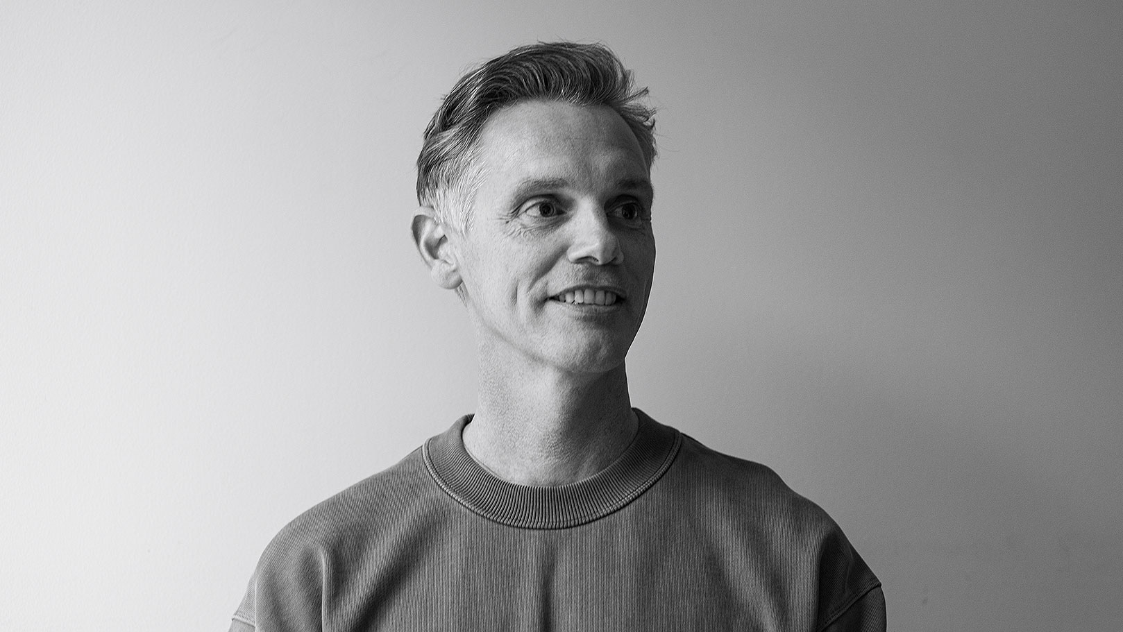 A black-and-white photo of Adrian Lawrence, Heckler's newly appointed Global ECD, with short, light hair, wearing a crew-neck sweatshirt, looking slightly to the side and smiling against a plain, light-colored background. Frame from Stash Magazine article.