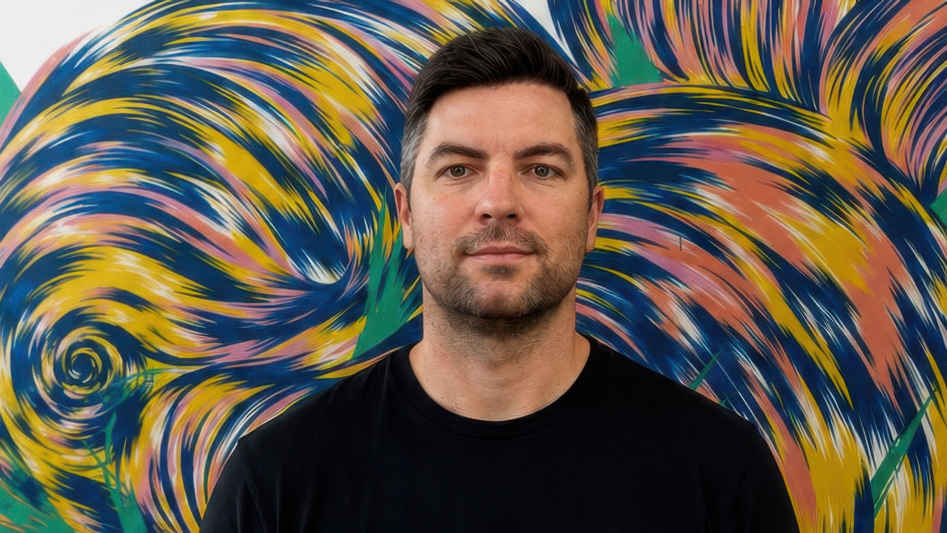 Heckler Singapore’s new Managing Director Jonathan Gerard, a man with short brown hair and a trimmed beard, stands in front of a colorful, abstract mural with swirling patterns in blue, yellow, pink, green, and orange. He wears a black t-shirt and looks directly at the camera. Frame from Stash Magazine article.