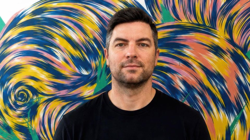 Heckler Singapore Managing Director Jonathan Gerard, a man with short dark hair and a trimmed beard, stands in front of a colorful, swirling abstract mural. He is wearing a plain black t-shirt and looks directly at the camera with a neutral expression. Frame from Stash Magazine article.