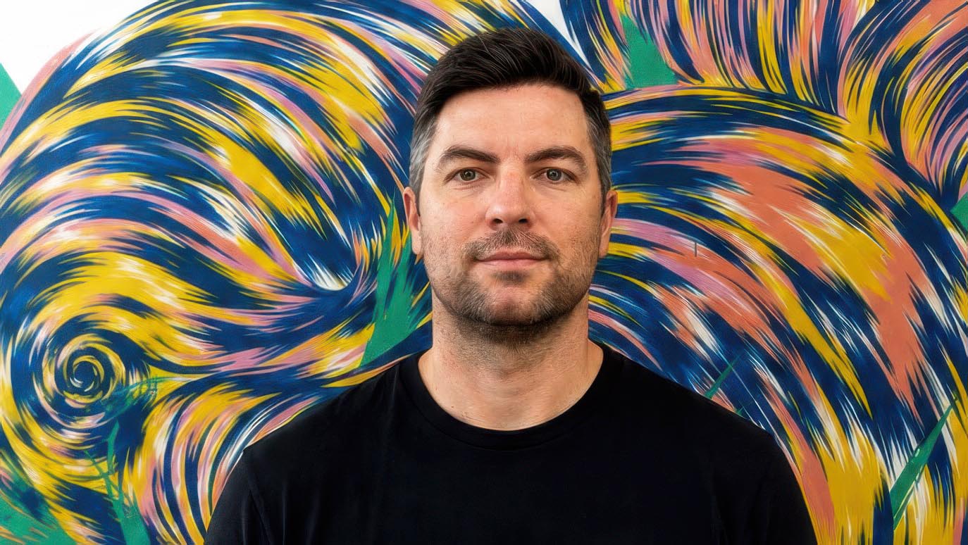 Heckler Singapore Managing Director Jonathan Gerard, a man with short dark hair and a trimmed beard, stands in front of a colorful, swirling abstract mural. He is wearing a plain black t-shirt and looks directly at the camera with a neutral expression. Frame from Stash Magazine article.