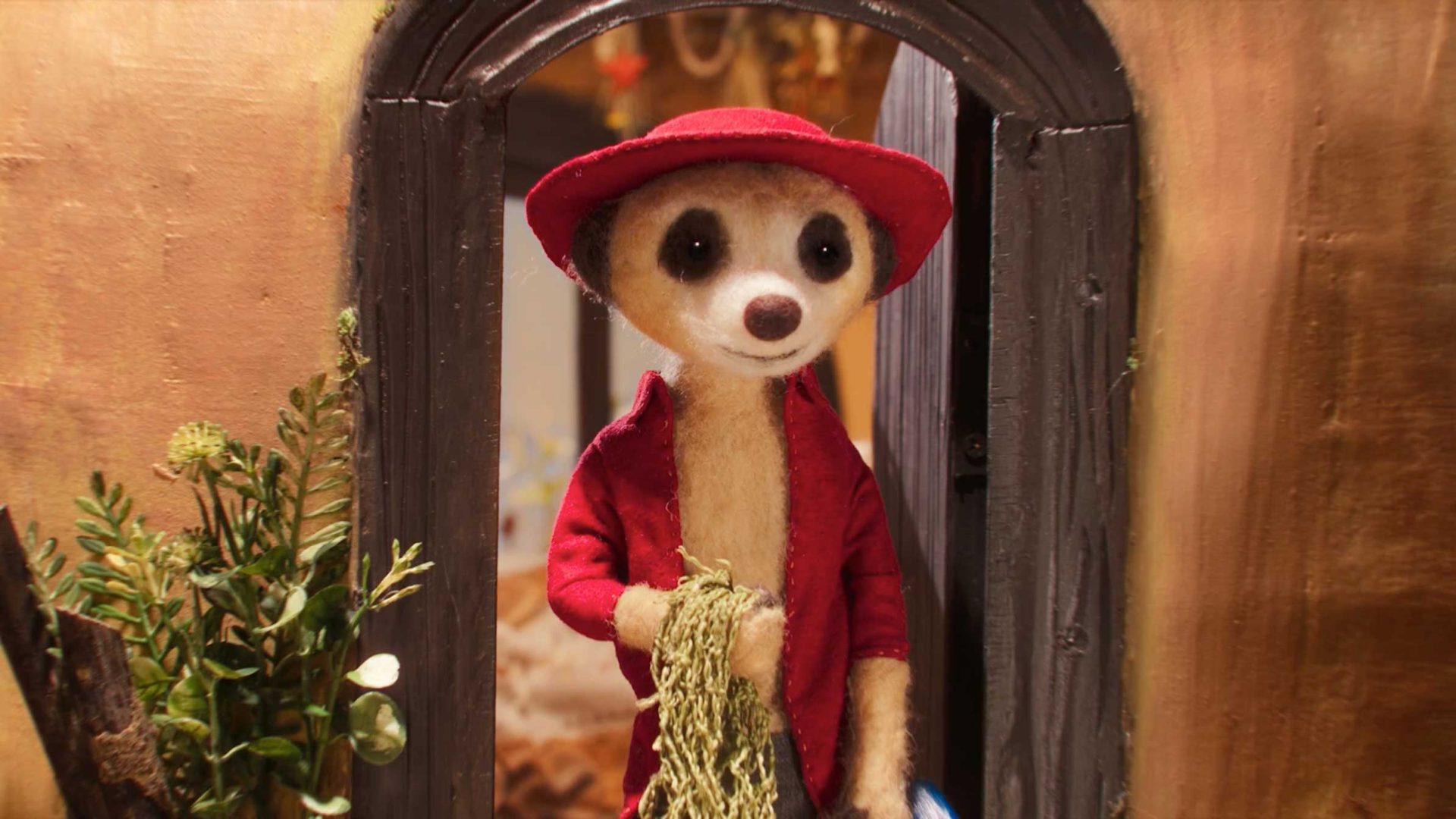 A plush meerkat in a red jacket and matching hat stands in a doorway, holding a bundle of hay like a whimsical Storyteller ready to spin a tale. Plants decorate the area around the entrance. Frame from Stash Magazine article.