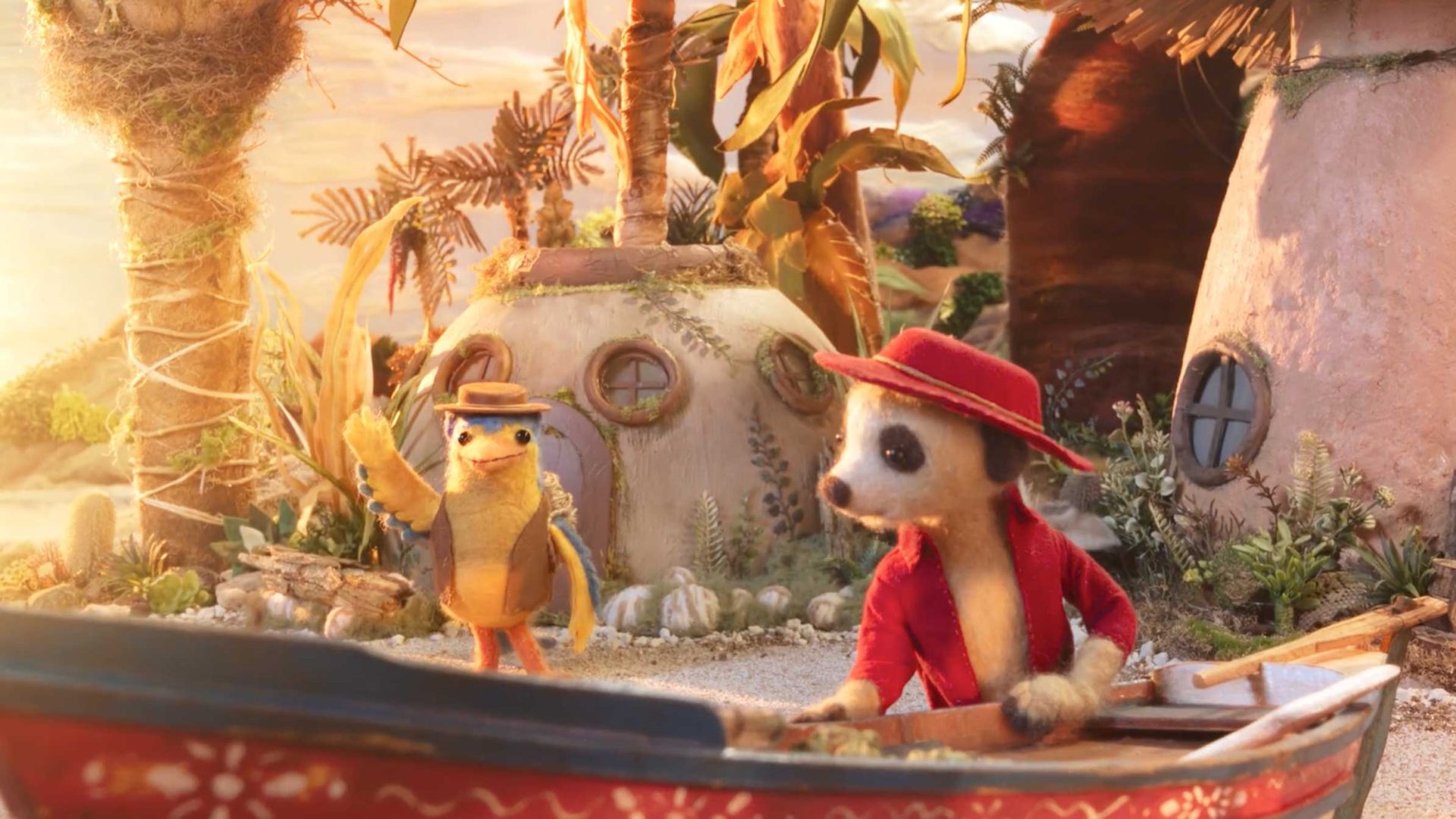 A meerkat in a red jacket and hat rows a boat on a sandy shore while a yellow lizard, known as the Jungler and also wearing a hat and jacket, waves nearby. Round, thatched-roof houses and tropical plants are in the background at sunset. Frame from Stash Magazine article.