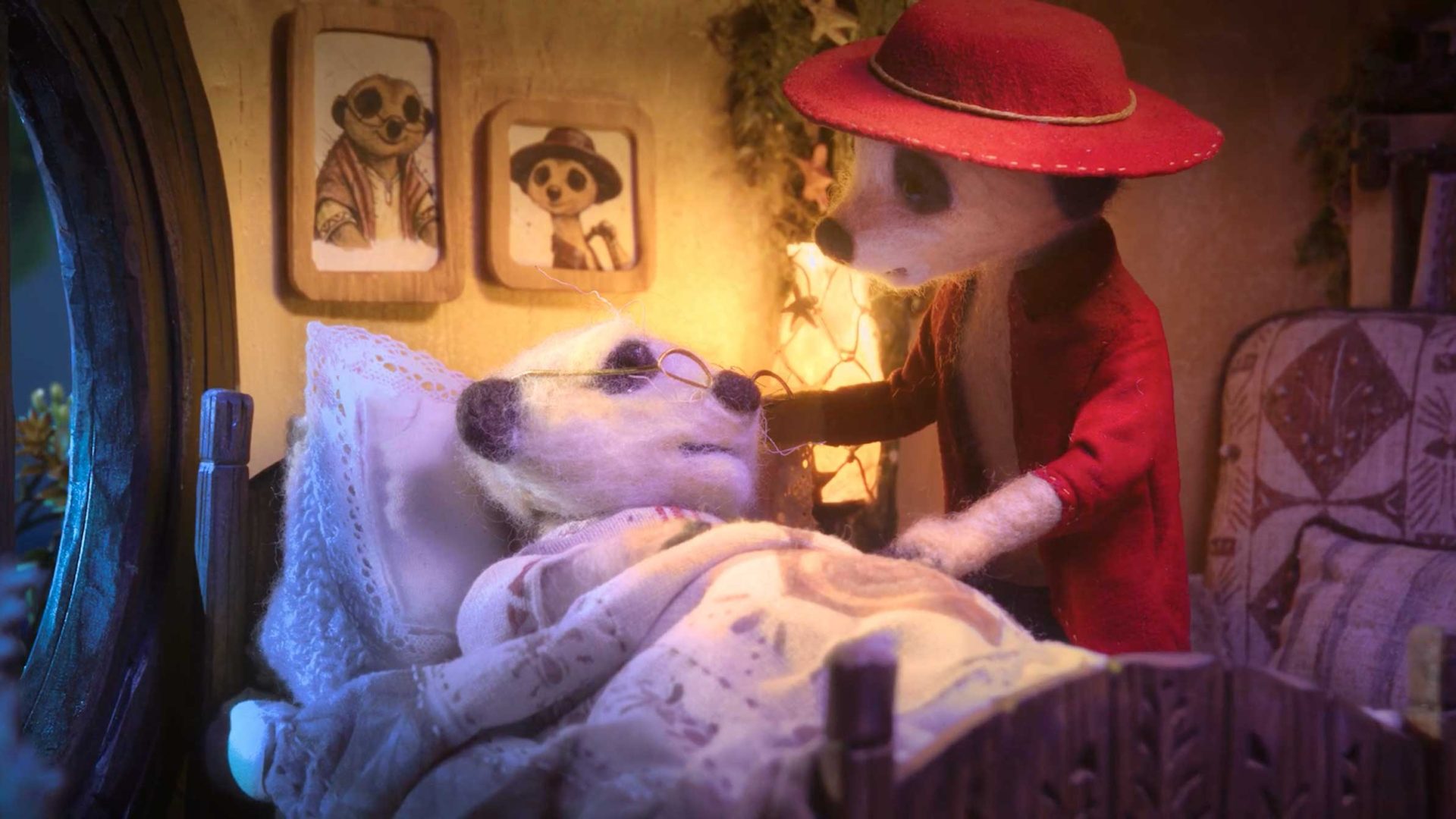 A stop-motion scene of two anthropomorphic meerkats: one lying in bed under a blanket wearing glasses, the other standing beside them in a red coat and hat, with warm lighting and framed portraits on the wall—much like how Zombie & Jungler told the story of The Storyteller for AXA, capturing both charm and heartfelt narrative. Frame from Stash Magazine article.