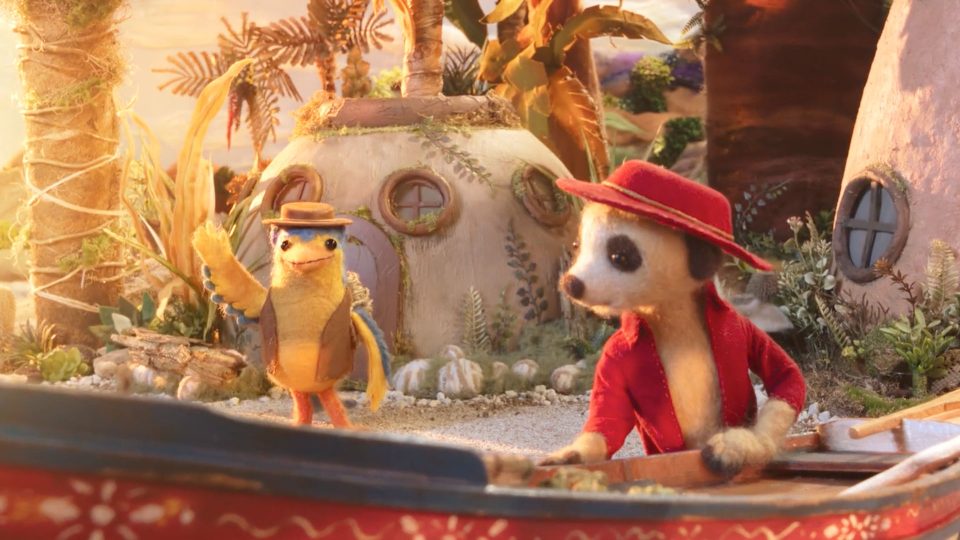 A meerkat in a red jacket and hat rows a boat, while a yellow bird in a hat waves nearby. In the background, The Storyteller’s round house with circular windows nestles among lush plants and trees, setting the scene for another whimsical adventure. Frame from Stash Magazine article.