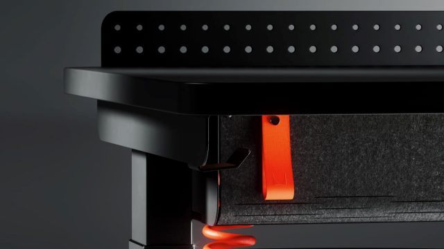Close-up of a modern black desk with perforated back panel, dark felt drawer, and an orange strap handle; a nod to Herman Miller Gaming style, with an orange cable visible underneath against a dark background. Frame from Stash Magazine article.