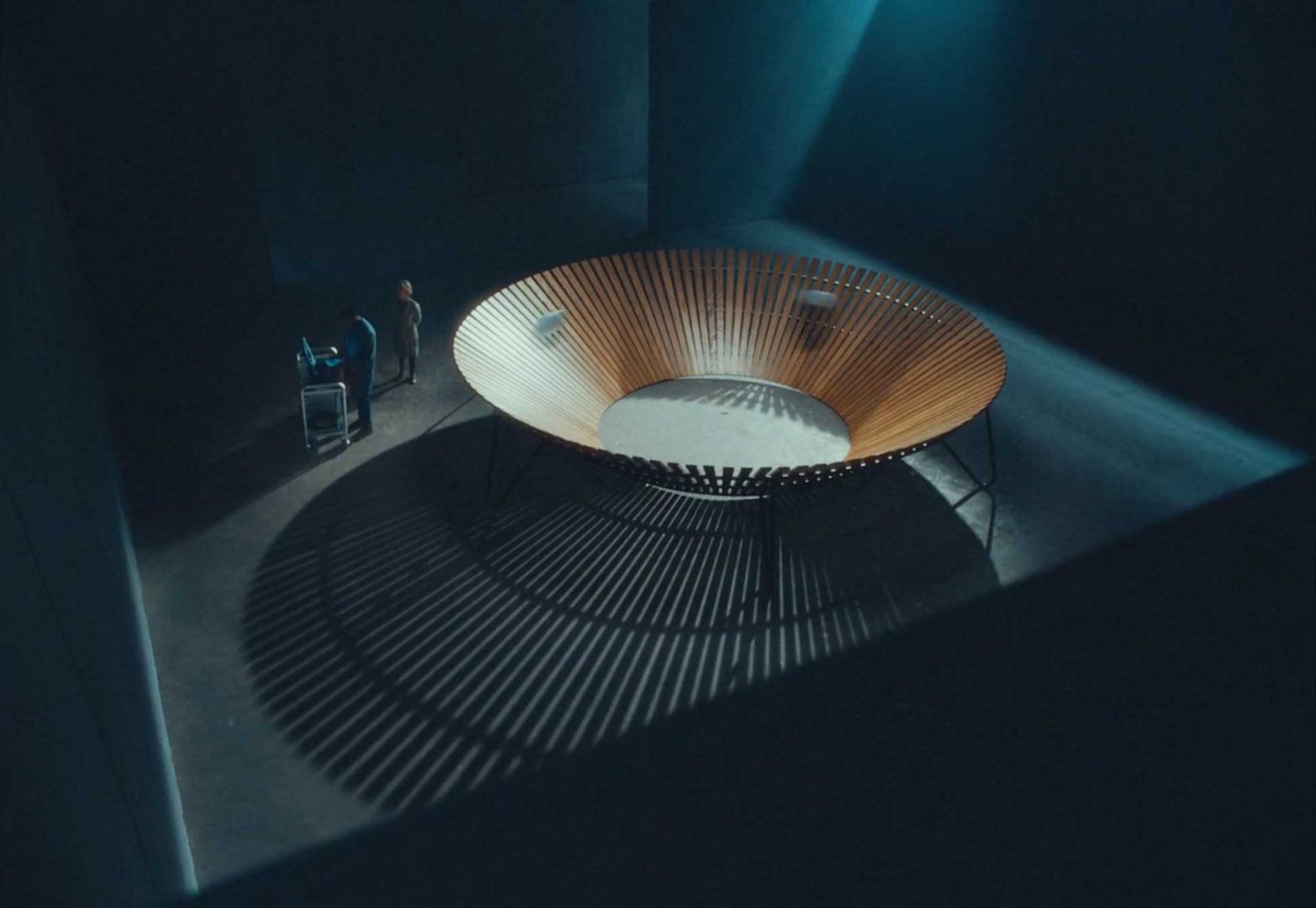 Two people stand near a large, circular, wooden structure with vertical slats, creating dramatic shadows on the floor in a dimly lit, spacious room—a setting where Dreams Engineered come to life in every detail. Frame from Stash Magazine article.