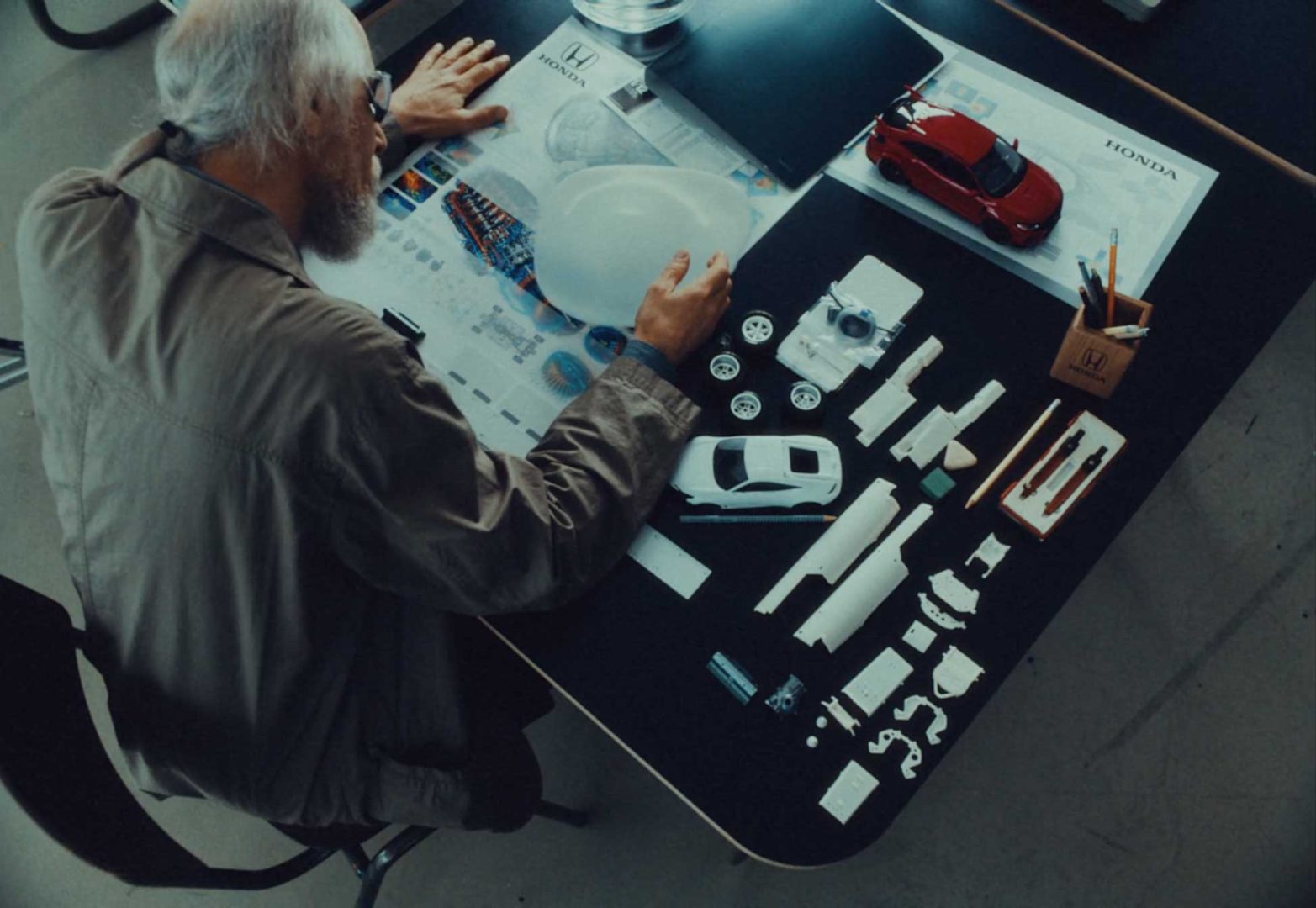An older man arranges car model parts and design plans on a table, including a red model car, Honda diagrams, and various white components, viewed from above—bringing to life the spirit of "Your Dreams Engineered. Frame from Stash Magazine article.