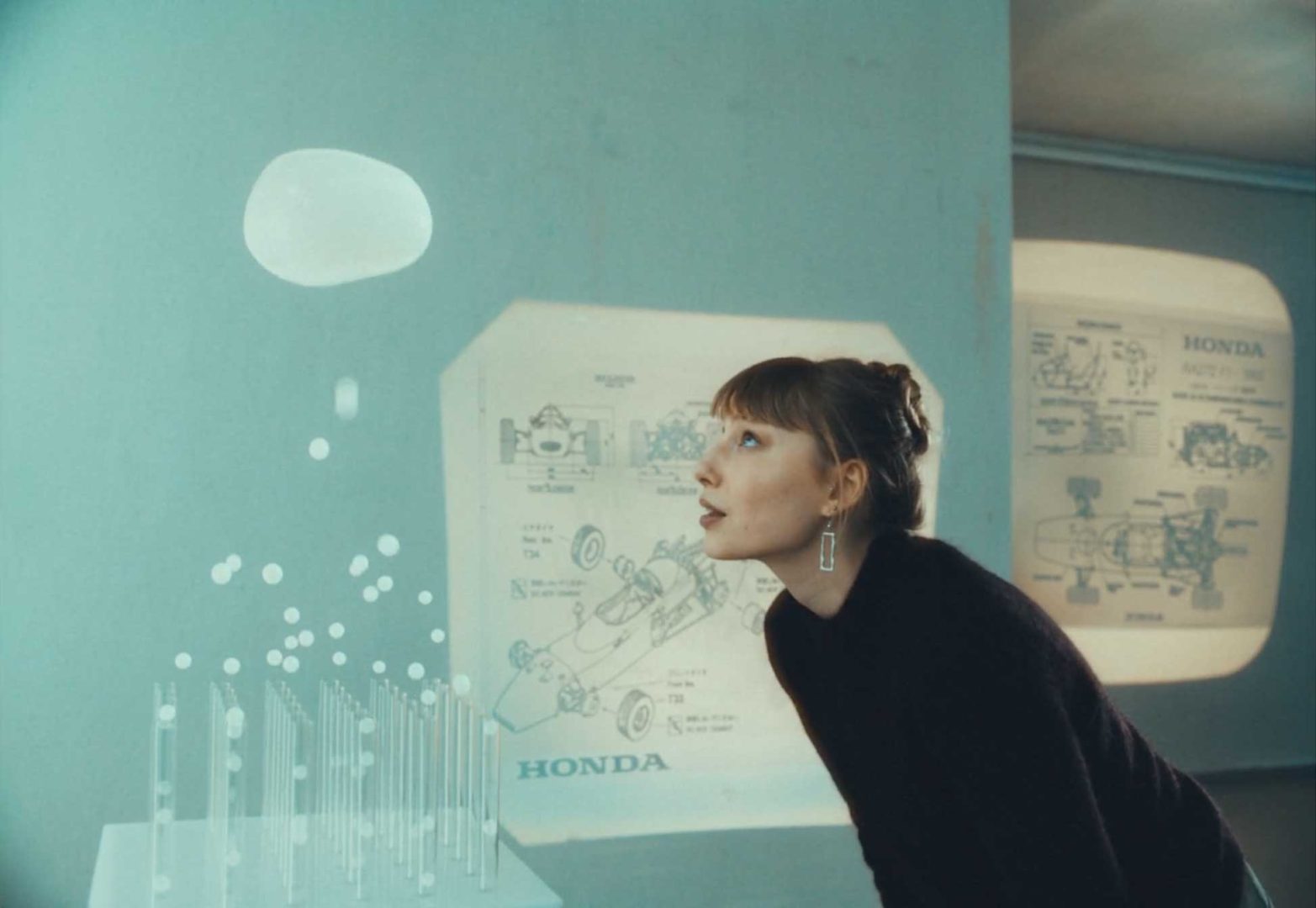 A woman looks at a floating translucent shape in a futuristic room with Honda engineering diagrams projected on walls, embodying the spirit of “Your Dreams Engineered” by Honda. Frame from Stash Magazine article.
