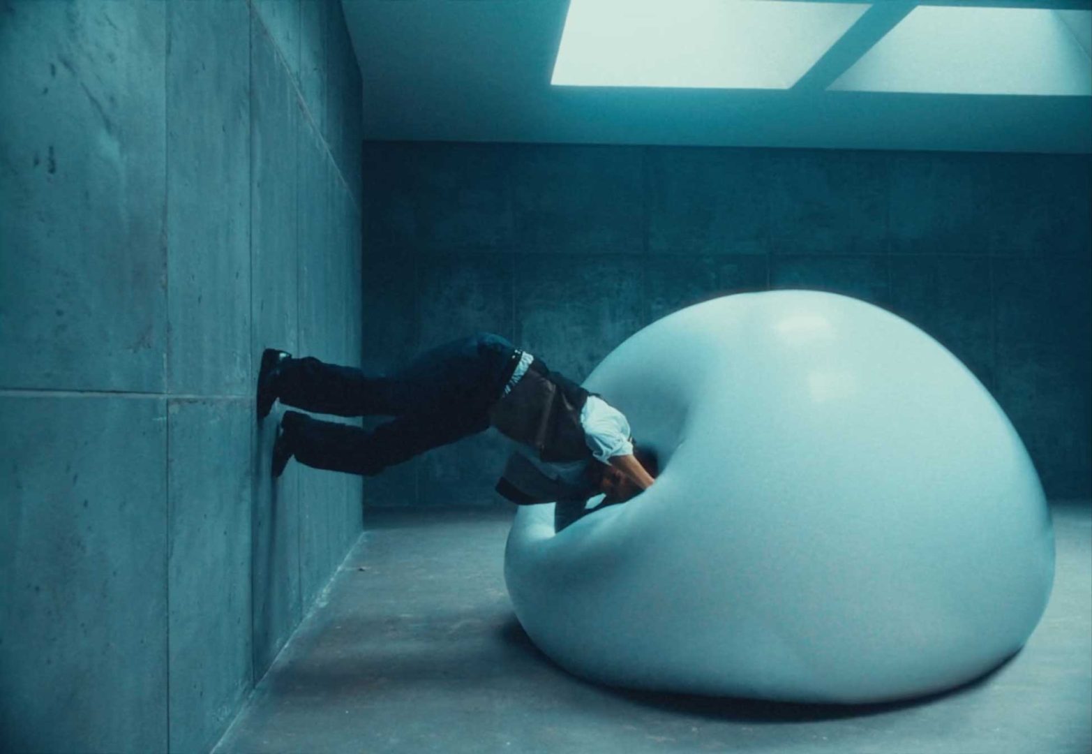 A person leans into a large, shiny, white blob against a concrete wall in a minimalist, blue-lit room with a skylight above—a futuristic scene where Dreams Engineered by Honda come to life. Frame from Stash Magazine article.