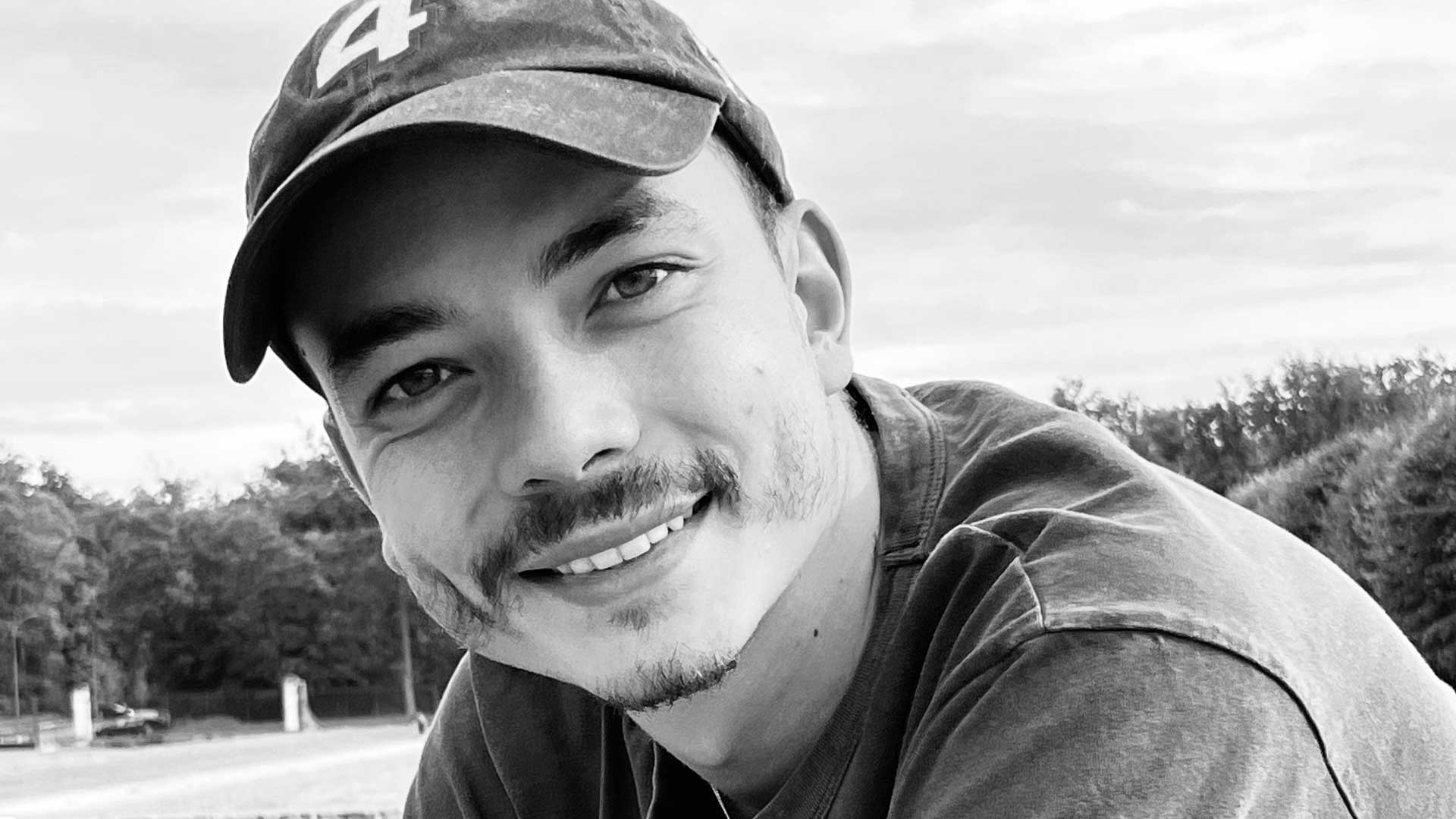 Hornet director Leo-Brunel, wearing a baseball cap and t-shirt, smiles at the camera outdoors. The black and white photo captures him against a backdrop of trees and a cloudy sky. Frame from Stash Magazine article.