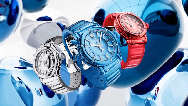 Three modern wristwatches—silver, blue, and red—are displayed floating in front of shiny metallic blue abstract shapes. Drawing inspiration from the Hublot x Jeff Koons Classic Fusion Balloon Dog by Zünc Studio, the background is bright, and the watches are prominently featured in the center. Frame from Stash Magazine article.