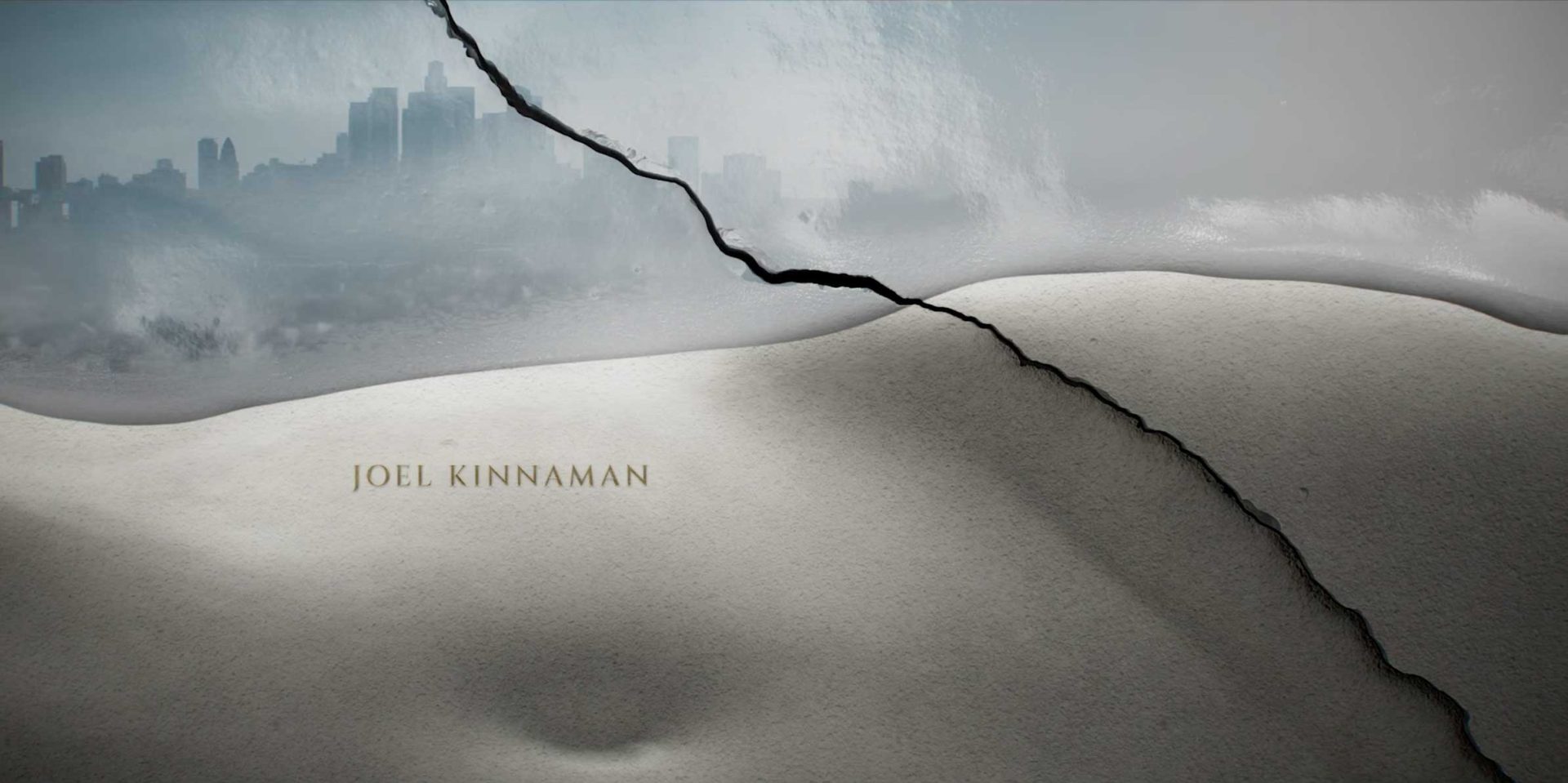 A dramatic crack splits a barren, sand-like landscape, with a hazy city skyline in the distance under a cloudy sky. Designed by Karin Fong of Imaginary Forces, the text JOEL KINNAMAN appears in gold lettering on the foreground. Frame from Stash Magazine article.