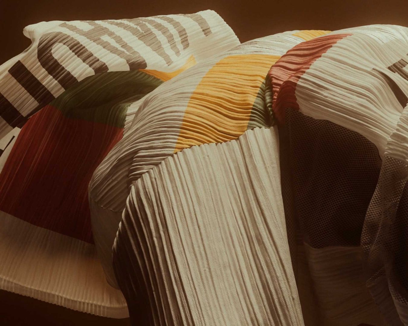 Close-up of pleated, textured fabric with colorful abstract patterns in white, yellow, green, and red tones, draped organically against a warm brown background, reminiscent of textiles seen in the Issey Miyake 1992 short film by Anatolik Belikov Studio. Frame from Stash Magazine article.