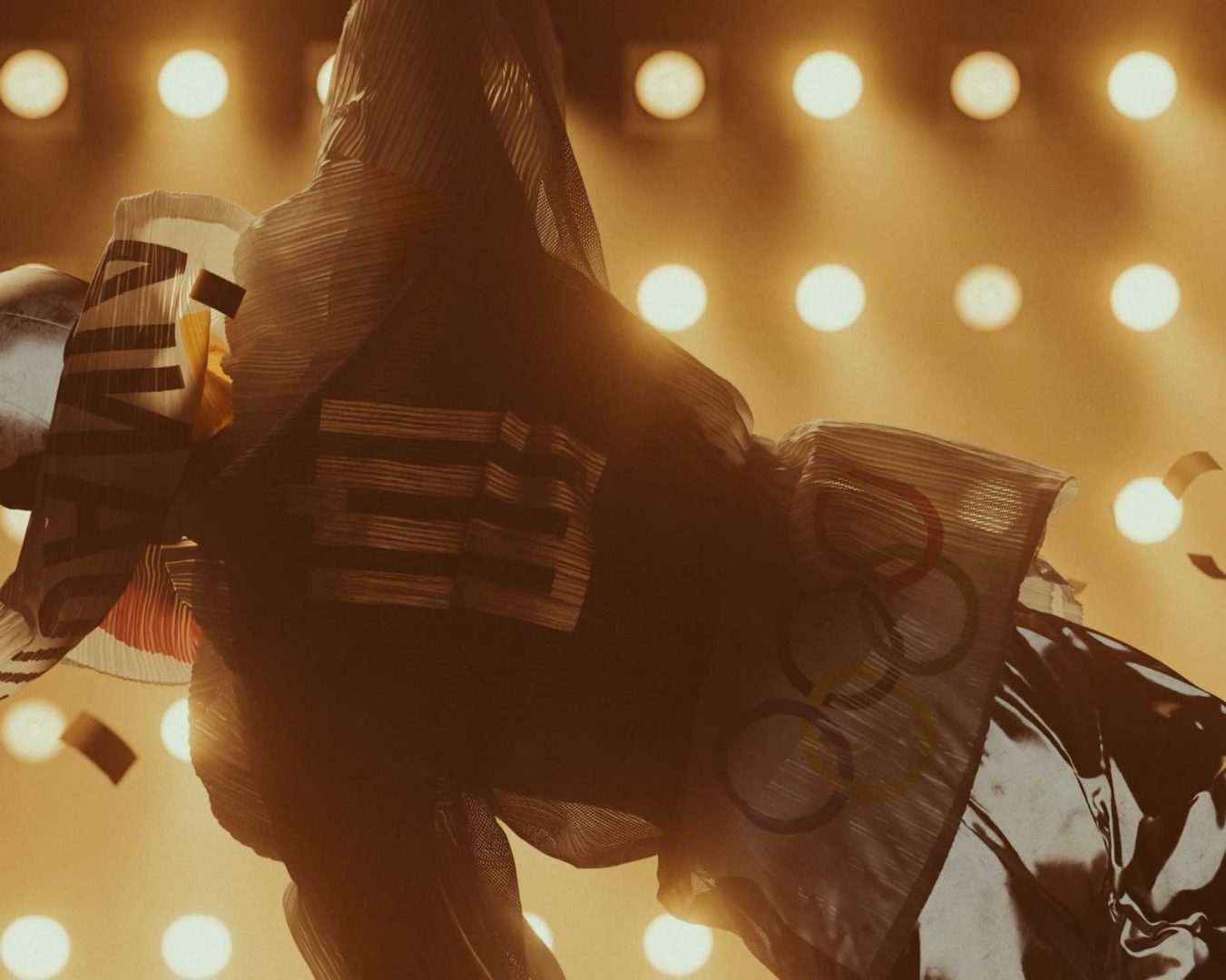 A person in a translucent jacket with DAVID and the Olympic rings stands under bright stage lights, confetti falling around them—a scene reminiscent of the avant-garde style seen in the Issey Miyake 1992 short film by Anatolik Belikov Studio. Frame from Stash Magazine article.