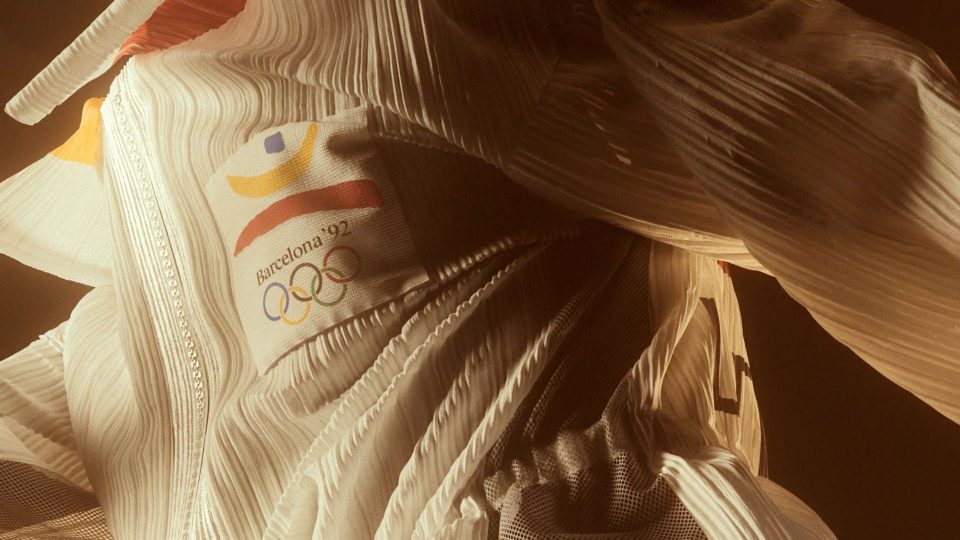 Close-up of a flowing, textured garment—echoing the spirit of Freedom Fashion—featuring the Barcelona ’92 Olympic Games logo and abstract shapes in red, yellow, and blue. The lighting and folds highlight the fabric’s details and movement, reminiscent of Issey Miyake’s innovative approach to textile design. Frame from Stash Magazine article.