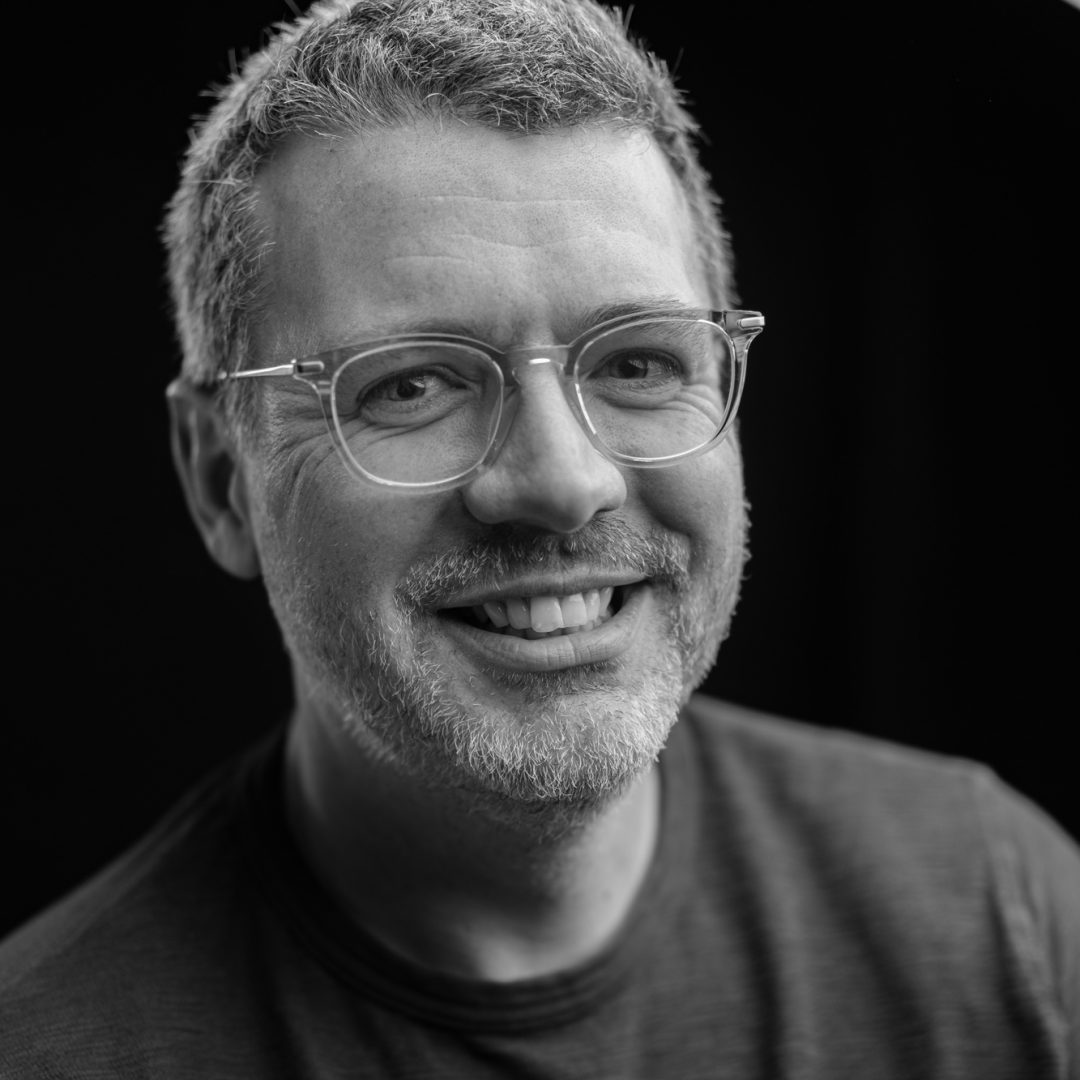 A man with short gray hair, glasses, and a beard smiles warmly at the camera. Wearing a t-shirt, he stands against a black background that gives the portrait a soft, professional look—perfect for OFFF 2026 or your Stash Panel to Get Seen. Frame from Stash Magazine article.