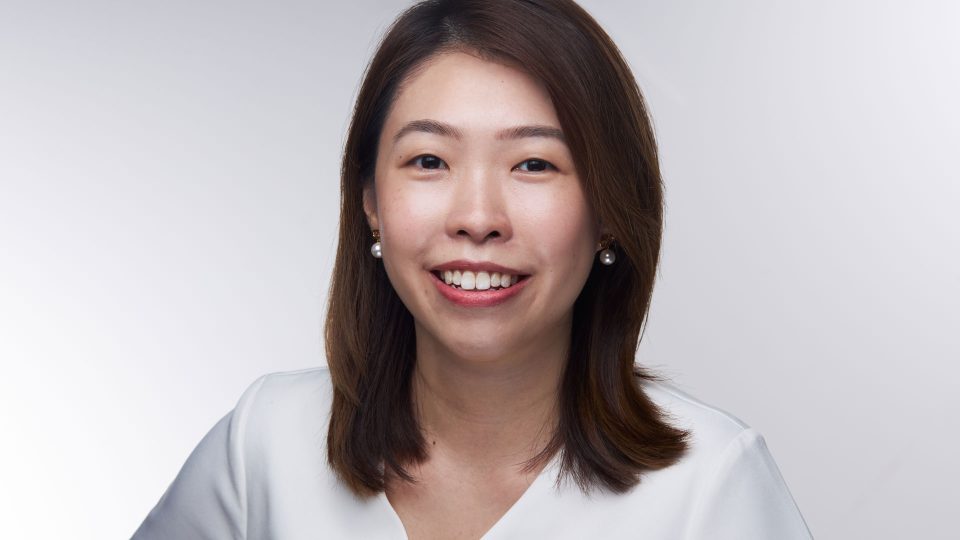 A woman with straight brown hair, wearing a white top and pearl earrings, smiles at the camera against a plain, light gray background—capturing the poised confidence reminiscent of Jasmine Tay, who was recently promoted to Executive Producer at Heckler Singapore. Frame from Stash Magazine article.