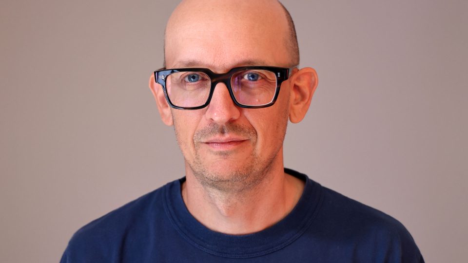Jim Allen, Executive WPP Creative Director, is pictured here with glasses, a shaved head, and a slight smile. He wears a navy blue shirt and looks directly at the camera against a plain light background—a classic portrait of one of the UK post production industry’s leading figures. Frame from Stash Magazine article.