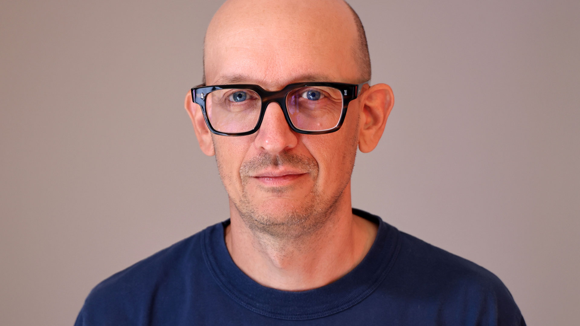 Jim Allen, Executive WPP Creative Director, is pictured here with glasses, a shaved head, and a slight smile. He wears a navy blue shirt and looks directly at the camera against a plain light background—a classic portrait of one of the UK post production industry’s leading figures. Frame from Stash Magazine article.