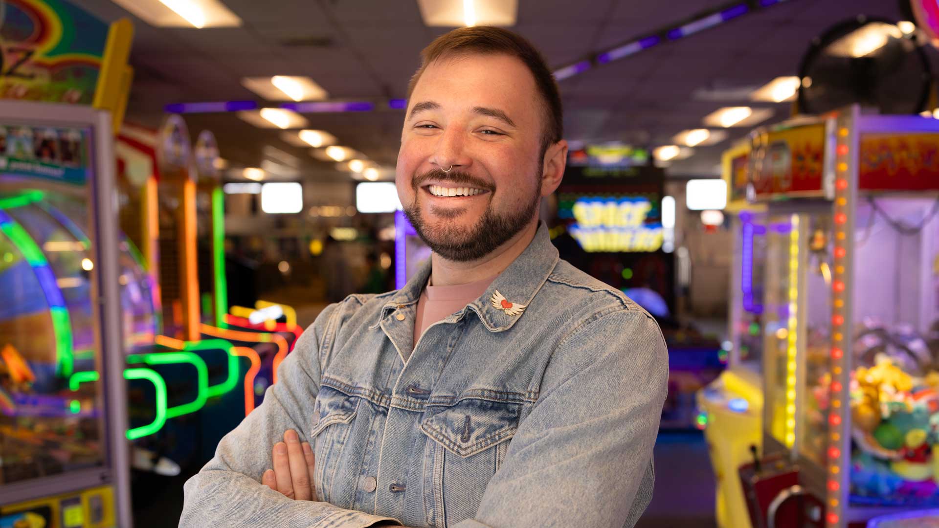 Joe Skoniecki, a Motion Graphics Designer, stands smiling with arms crossed in a denim jacket at a brightly lit arcade, surrounded by colorful games and machines. Frame from Stash Magazine article.