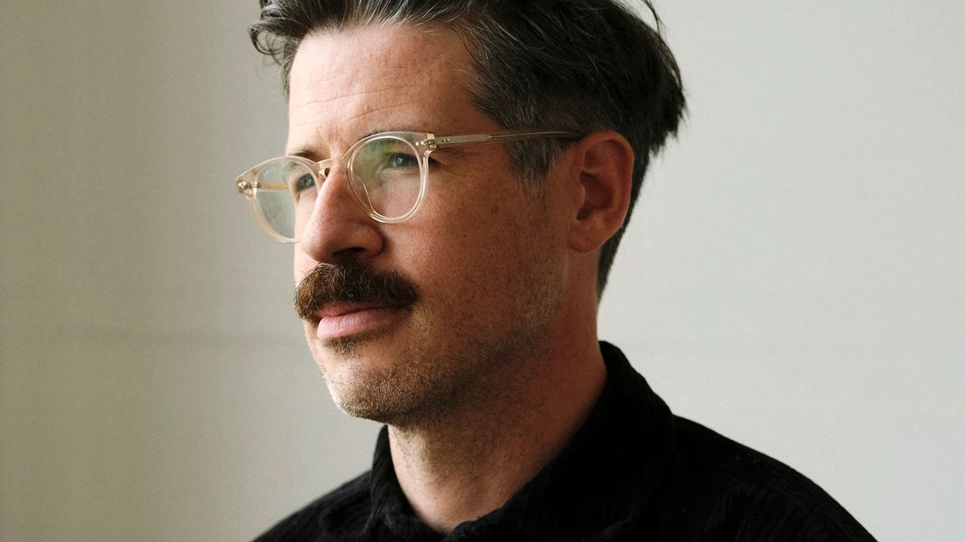 A man with short dark hair, a mustache, and light beard wears clear glasses and a black shirt, looking thoughtfully to the side against a plain light background—a scene reminiscent of Johnny-Kelly-Signs-to-Riff-Raff. Frame from Stash Magazine article.