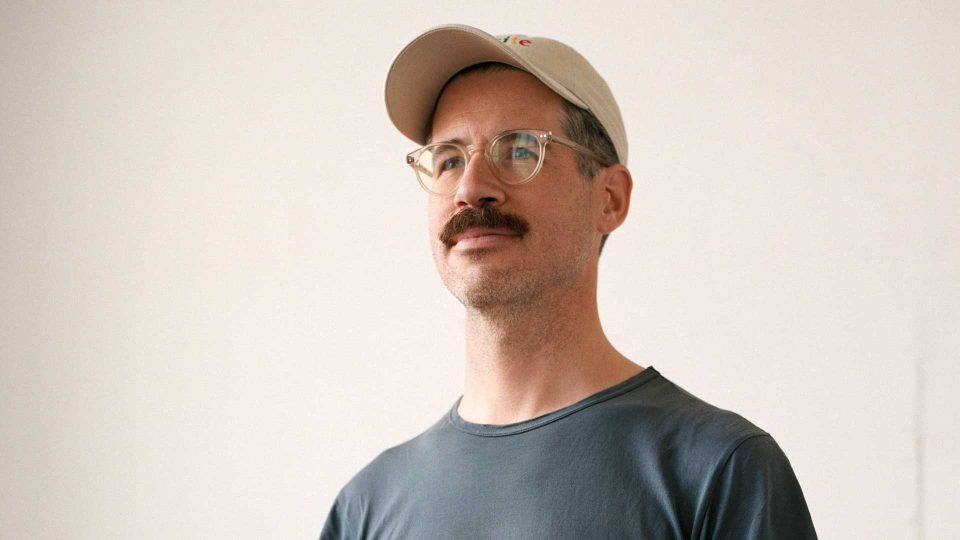 A man with a mustache and glasses wears a beige baseball cap and a dark t-shirt, standing against a plain light-colored background, evoking the style seen in Johnny-Kelly-Signs-to-Riff-Raff. Frame from Stash Magazine article.