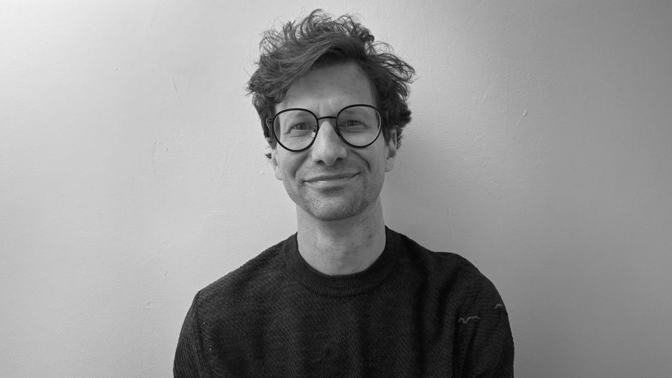 A person with curly hair, wearing round glasses and a dark textured sweater, smiles slightly while sitting against a plain, light-colored wall. The black-and-white photo features Jon Lerner, Head of Experiential at Versus. Frame from Stash Magazine article.