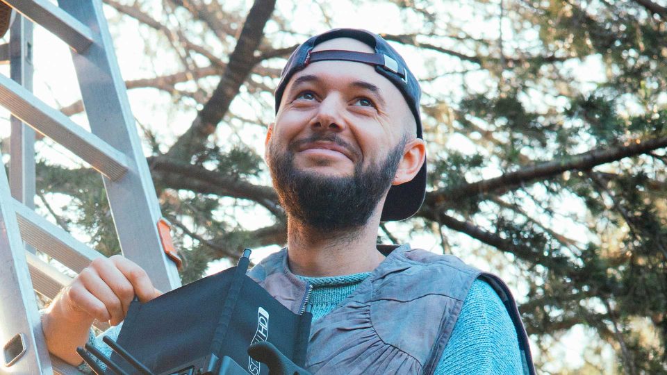 A bearded person wearing a backward cap smiles while standing outdoors near a metal ladder, holding a black object. Sunlight filters through tree branches in the background, evoking the creative spirit that Joseph Mann brings as he signs with Caviar for US commercial representation. Frame from Stash Magazine article.