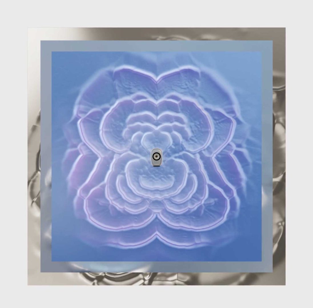 A square artwork with a soft, abstract blue and violet floral pattern radiates from a small black-and-white circle at the center, bordered by a metallic silver frame. Reminiscent of the refined aesthetic found in James Brocklebank’s LS Luxe collection for KEF, the background is a light gray. Frame from Stash Magazine article.
