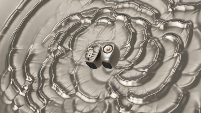 Two wireless earbuds rest on a rippled, metallic surface with concentric circular patterns spreading outward, creating a sense of sound waves—a visual reminiscent of the artistry seen in the KEF LS Luxe x Ross Lovegrove launch film by James Brocklebank. Frame from Stash Magazine article.