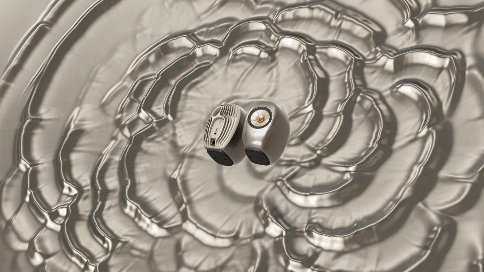 Two wireless earbuds rest on a rippled, metallic surface with concentric circular patterns spreading outward, creating a sense of sound waves—a visual reminiscent of the artistry seen in the KEF LS Luxe x Ross Lovegrove launch film by James Brocklebank. Frame from Stash Magazine article.