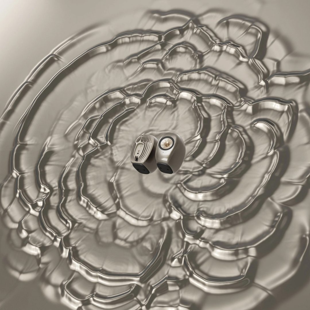 Two small KEF speakers sit side by side on a light, reflective surface, surrounded by rippling wave patterns that radiate outward, suggesting the movement of sound—an effect reminiscent of the acoustic artistry envisioned by James Brocklebank in the LS Luxe series. Frame from Stash Magazine article.