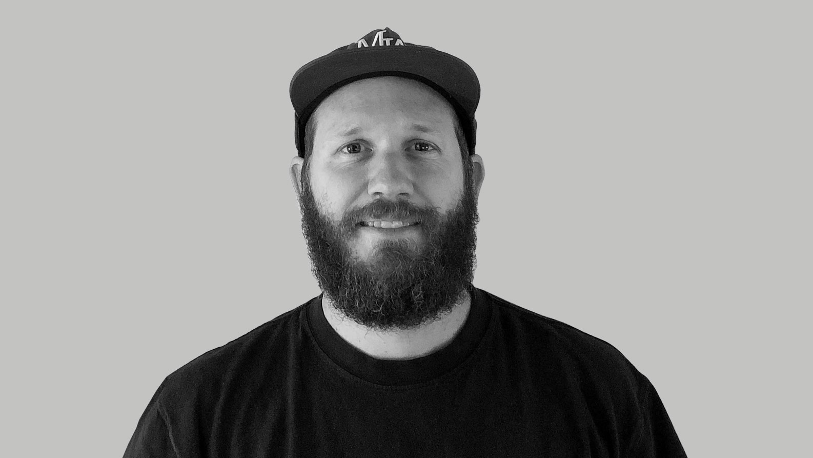 A man with a full beard wearing a black t-shirt and a baseball cap, smiling at the camera against a plain grey background, celebrates as Kev Jones is named Executive Producer at Scholar. Frame from Stash Magazine article.