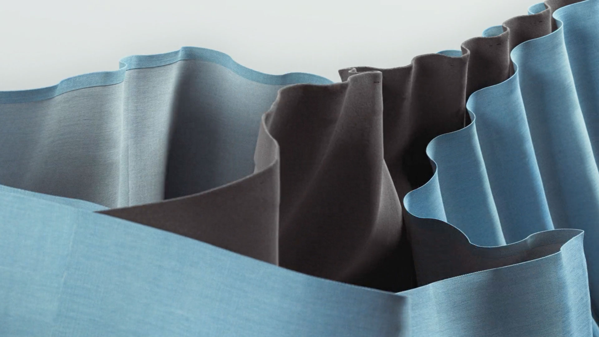 Close-up of wavy, layered Kvadrat fabric in shades of blue and gray, arranged in soft folds against a light background. The textures and curves create an abstract pattern reminiscent of gentle waves. Frame from Stash Magazine article.