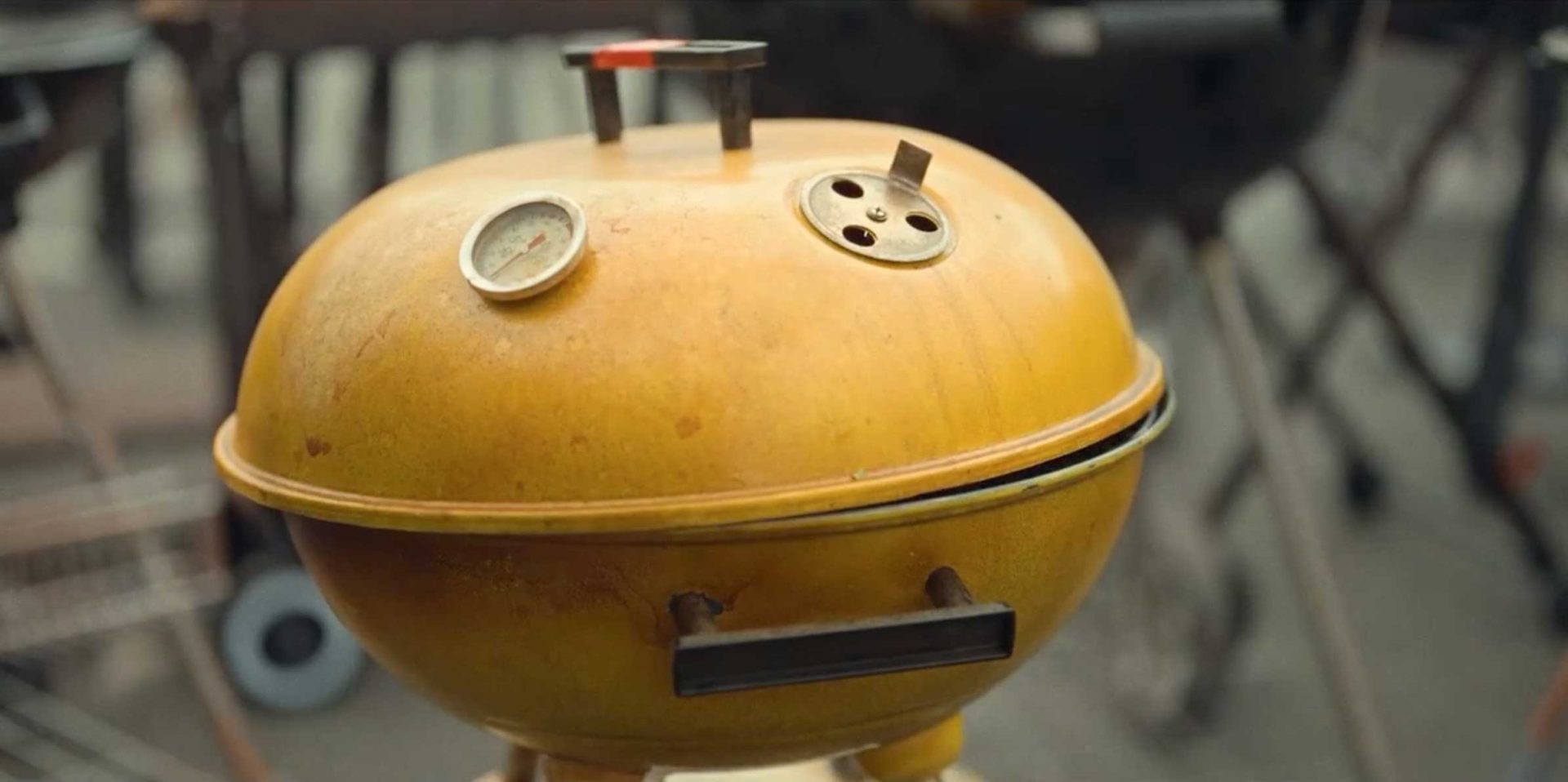 A yellow round charcoal grill with a thermometer and vent holes on top, resembling a face with two eyes and a rectangular mouth-like handle, is shown outdoors—perfect for channeling your inner Barbecue Piper at any Lidl Finland backyard cookout. Frame from Stash Magazine article.